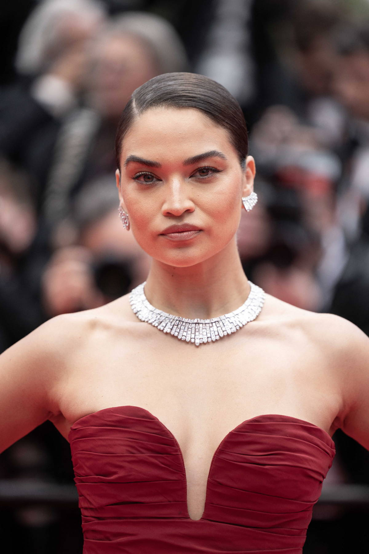 Woman with slicked-back hair wearing a strapless deep red gown with a plunging neckline, accessorized with a sparkling necklace and matching earrings, standing confidently on the red carpet with a serious expression and a blurred background of a formal event