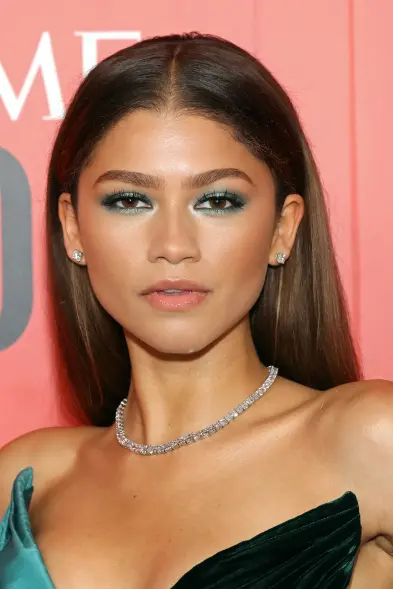 Elegant woman with long, sleek hair and striking blue eyeshadow, wearing diamond stud earrings and a diamond necklace, posing against a red background.