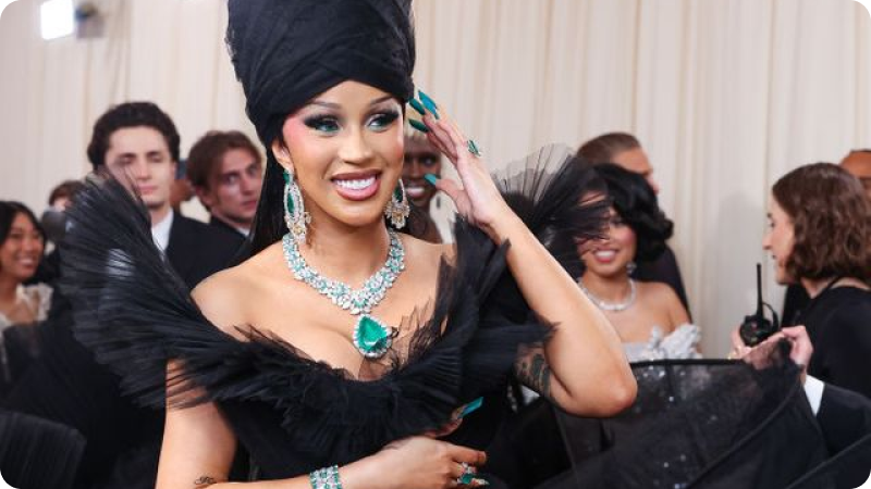 A woman dressed in an elaborate black gown with dramatic ruffled details and a large matching headpiece is adorned with sparkling jewelry, including a striking emerald necklace, earrings, and rings. She is smiling brightly, showing vibrant makeup and long, turquoise nails. The background features other event attendees, adding to the festive and glamorous atmosphere.