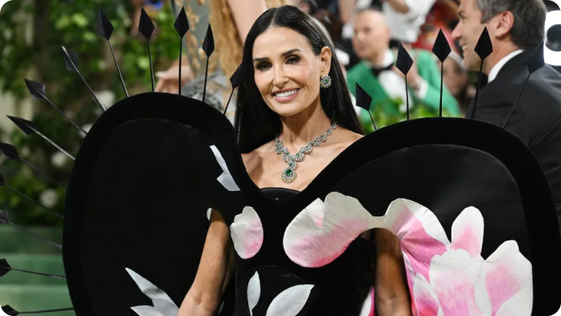 A woman with long, dark hair is wearing a dramatic, avant-garde black gown with large, butterfly-like wings featuring pink and white floral patterns. She is adorned with an elegant necklace and earrings that feature green gemstones. She is smiling confidently, and the background shows other event attendees and a lush, green setting, highlighting the glamorous atmosphere.