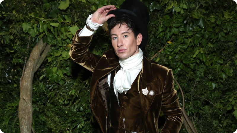 A man dressed in an elaborate, vintage-inspired outfit featuring a velvet coat, a white cravat, and a top hat. He is posing with one hand holding the brim of his hat. The background consists of lush green foliage, creating a dramatic and elegant atmosphere.