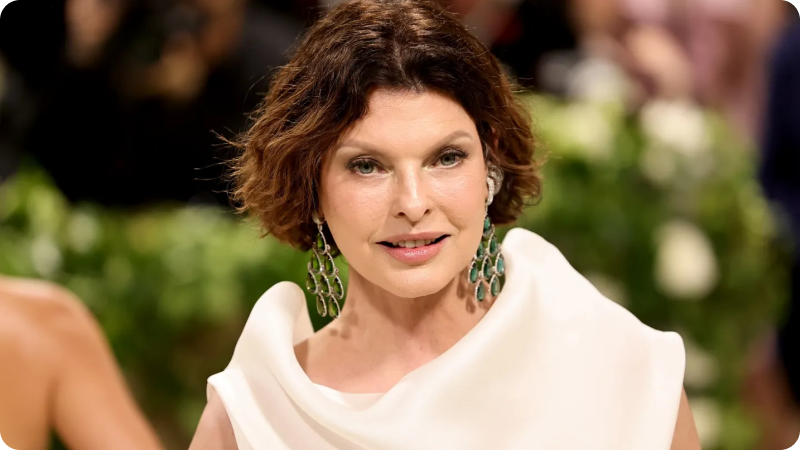 A woman with short, wavy brown hair is wearing a sophisticated white gown with a draped neckline. She is accessorized with striking green chandelier earrings. The background features a lush green setting and blurred event attendees, emphasizing the elegant and glamorous atmosphere of the event.