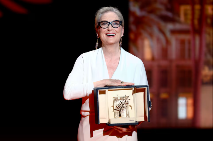 Woman in glasses and a white dress smiling on stage while holding the Palme d'Or award in a box, with a blurred background and warm lighting indicating a formal event.