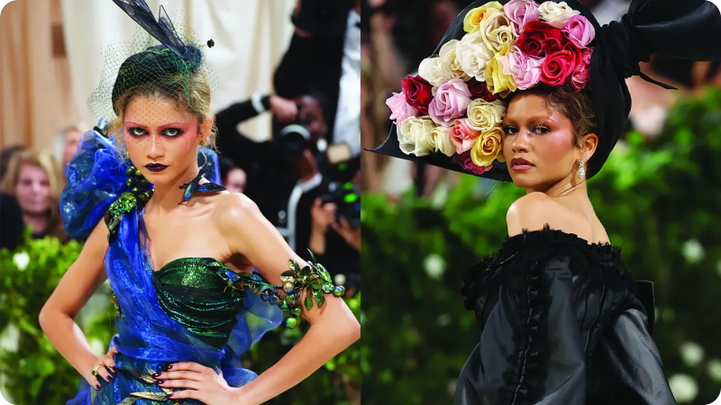 Two women at a glamorous event, each wearing distinctive and elaborate outfits. The woman on the left is dressed in a vibrant green and blue gown with intricate details, complemented by dramatic makeup and a feathered headpiece. The woman on the right is wearing a black outfit with a large, colorful floral headpiece, featuring roses in various shades. Both are posing confidently against a lush, green backdrop with photographers and onlookers in the background