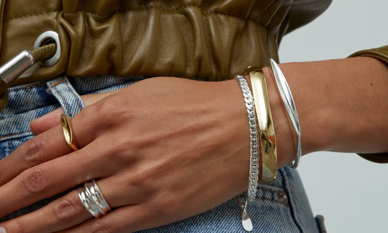 Close-up of a woman's hand adorned with multiple rings and bracelets, wearing a brown leather jacket and blue jeans.