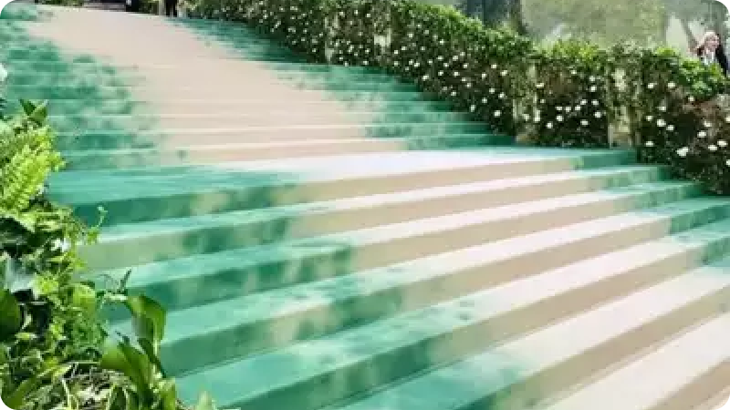The image shows a set of stairs with a green and white striped pattern, adorned with lush green foliage and white flowers along the sides, giving it a vibrant, garden-like appearance
