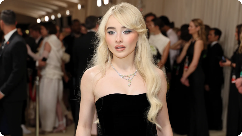 A woman with long, blonde hair is wearing a strapless black gown and a delicate necklace. She has soft makeup with a subtle, elegant look. The background features other event attendees and a well-lit, sophisticated setting, highlighting the glamorous atmosphere of the event.