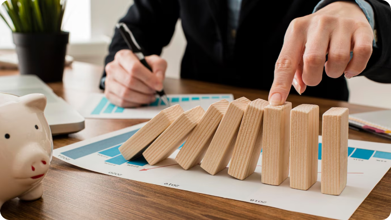 Person preventing wooden blocks from falling, symbolizing stability and risk management, with a piggy bank and financial charts on the desk in the background