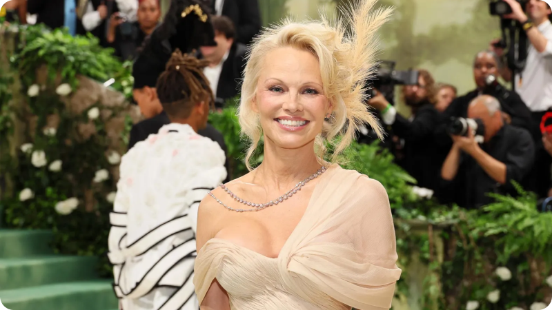 A woman with blonde hair styled in an elegant updo, adorned with feather details, is wearing a flowing, off-the-shoulder cream gown. She has a radiant smile and is accessorized with a delicate necklace. The background features a lush, green setting with photographers and other event attendees, highlighting the glamorous and sophisticated atmosphere of the event.