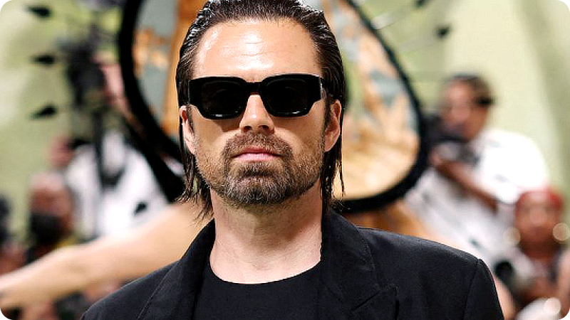 A man with slicked-back hair and a beard is wearing black sunglasses and a black outfit, including a blazer and a T-shirt. He has a serious expression. The background features a blurred crowd of photographers and other attendees, emphasizing the sophisticated and fashionable atmosphere of the event.