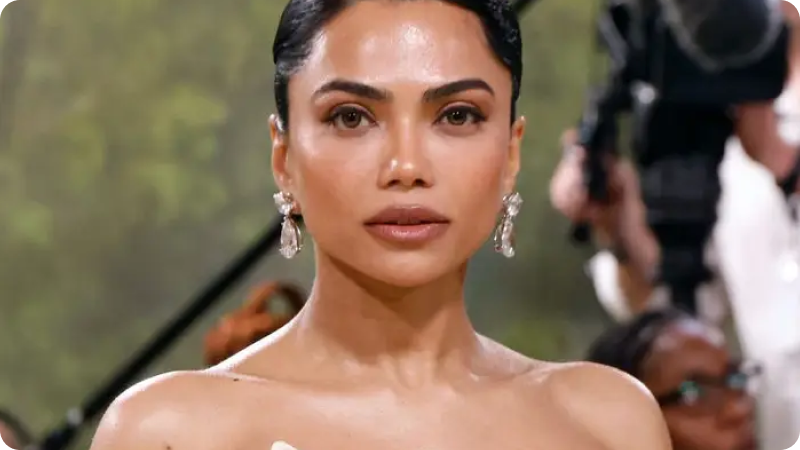 A woman with sleek, dark hair is wearing elegant, sparkling drop earrings and has a radiant, natural makeup look. She is dressed in a sophisticated off-the-shoulder gown. The background shows a blurred event setting with other attendees and photographers, highlighting the formal and glamorous atmosphere.